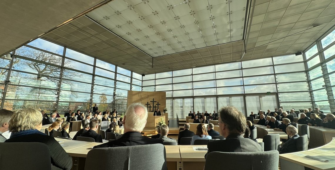 Blick in den hellen Plenarsaal des Schleswig-Holsteinischen Landtags. Zahlreiche Gäste sitzen in Reihen an Tischen, während vorne am Rednerpult der Landesvorsitzende des Volksbundes, Dr. Ekkehard Klug, spricht. Hinter ihm sind fünf Kreuze zu sehen. Große Glasfronten lassen viel Tageslicht in den Raum.