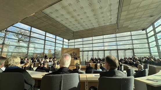 Blick in den hellen Plenarsaal des Schleswig-Holsteinischen Landtags. Zahlreiche Gäste sitzen in Reihen an Tischen, während vorne am Rednerpult der Landesvorsitzende des Volksbundes, Dr. Ekkehard Klug, spricht. Hinter ihm sind fünf Kreuze zu sehen. Große Glasfronten lassen viel Tageslicht in den Raum.