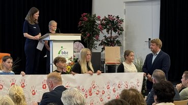 Ein Mädchen hält auf einem Podest eine Rede. Im Vordergrund sind weitere Kinder zu sehen, die ein weißes Banner mit Roten Handabdrücken hochhalten.
