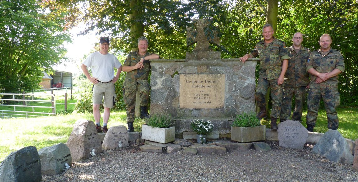 Fünf Personen stehen vor einem Gedenkstein im Freien. Vier tragen Tarnuniformen, eine Person ist in Freizeitkleidung mit weißem T-Shirt und khakifarbenen Shorts gekleidet. Der Gedenkstein ist von kleineren Steinen und Pflanzen umgeben, im Hintergrund sind Bäume und Grünflächen zu sehen.