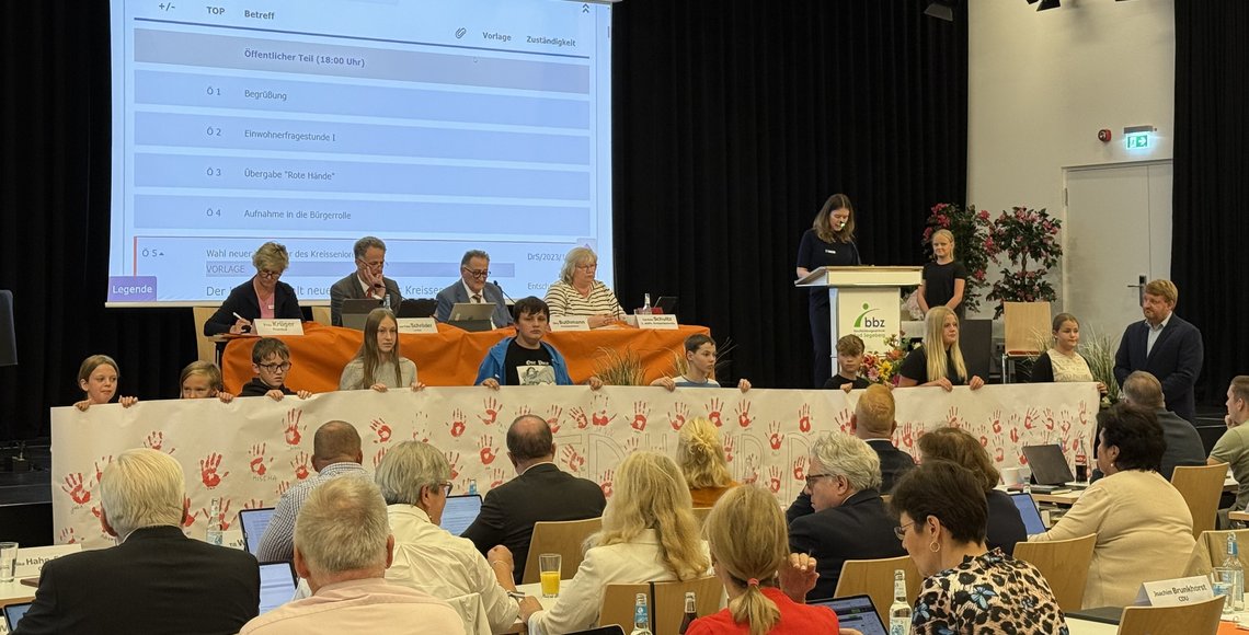 Eine Gruppe von Schülerinnen und Schülern präsentiert ein weißes Banner mit roten Handabdrücken. Im Hintergrund stehen Kreistagspolitiker, im Vordergrund sind weitere Kreistagsabgeordnete zu sehen.