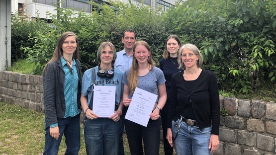 Sechs Personen stehen nebeneinander vor einem Schulgebäude. Keira Pahl und Finja Baaß halten ihre Urkunden zur Teilnahme am Internationalen Comic-Wettbewerb in den Händen