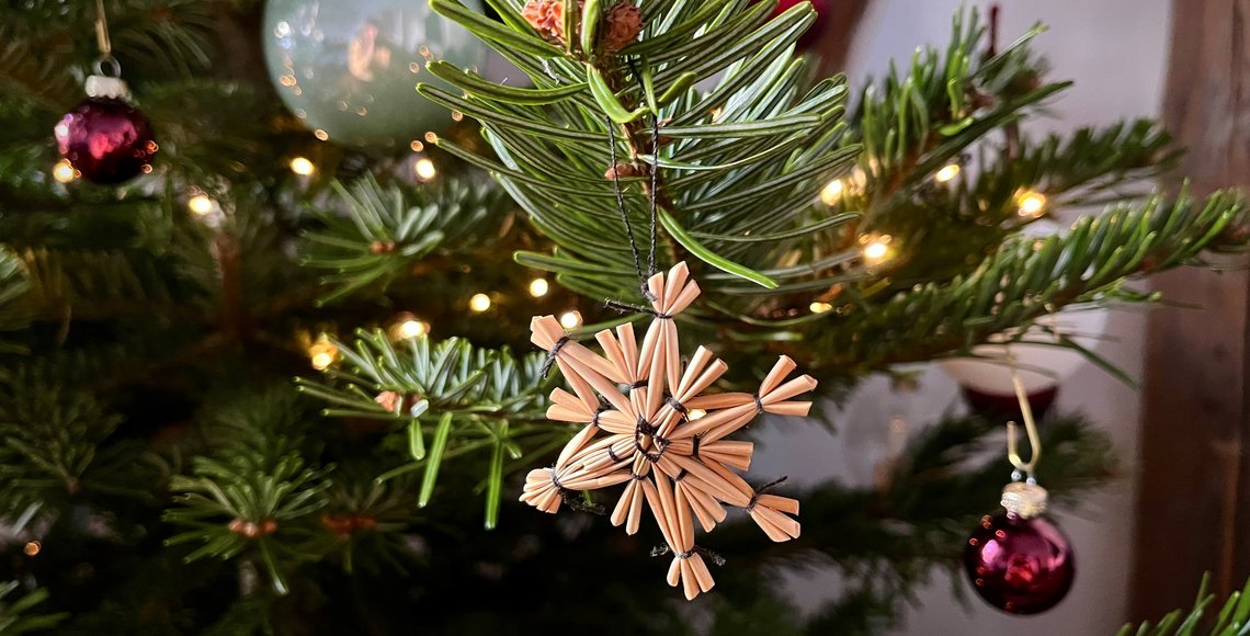 Nahaufnahme eines geschmückten Weihnachtsbaums mit grünem Tannenzweig im Vordergrund. Daran hängt ein handgefertigter Strohstern in Form einer Schneeflocke. Im Hintergrund sind kleine, warme Lichterketten und rote Christbaumkugeln zu sehen