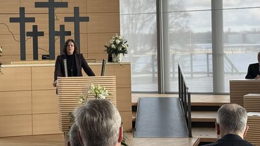 Rednerin steht am Pult im Plenarsaal des Schleswig-Holsteinischen Landtags. Hinter ihr eine große Holztafel mit einer stilisierten Landkarte von Schleswig-Holstein und fünf schwarzen Kreuzen als Symbol des Gedenkens. Links und rechts des Pults stehen Blumengestecke, im Hintergrund große Fensterfront mit Blick ins Freie