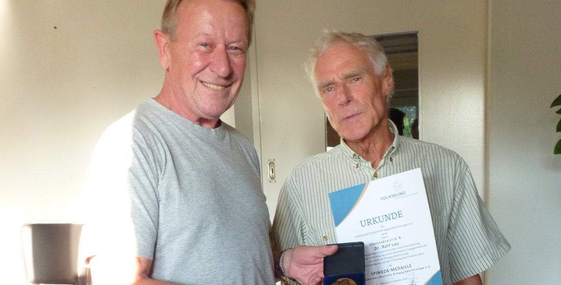 Zwei Männer stehen in einem Innenraum und lächeln in die Kamera. Die Person auf der linken Seite hält eine Medaille. Die Person auf der rechten Seite hält eine Urkunde..