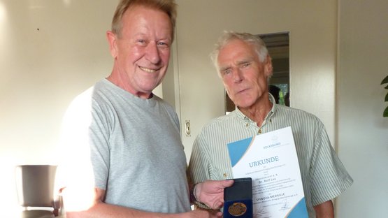 Zwei Männer stehen in einem Innenraum und lächeln in die Kamera. Die Person auf der linken Seite hält eine Medaille. Die Person auf der rechten Seite hält eine Urkunde..