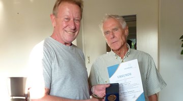 Zwei Männer stehen in einem Innenraum und lächeln in die Kamera. Die Person auf der linken Seite hält eine Medaille. Die Person auf der rechten Seite hält eine Urkunde..