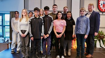 Auf dem Bild sind Schülerinnen und Schüler der Gemeinschaftsschule am Marschweg zusammen mit ihrem Lehrer auf einer Bühne zu sehen. Sie stehen nebeneinander und lächeln in die Kamera.