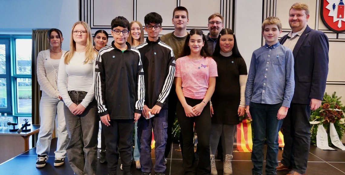 Auf dem Bild sind Schülerinnen und Schüler der Gemeinschaftsschule am Marschweg zusammen mit ihrem Lehrer auf einer Bühne zu sehen. Sie stehen nebeneinander und lächeln in die Kamera.