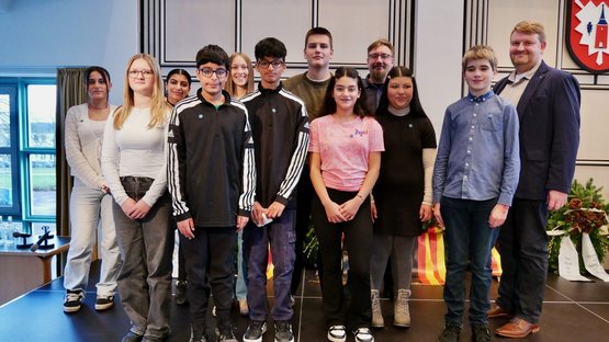 Auf dem Bild sind Schülerinnen und Schüler der Gemeinschaftsschule am Marschweg zusammen mit ihrem Lehrer auf einer Bühne zu sehen. Sie stehen nebeneinander und lächeln in die Kamera.