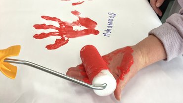 Zu sehen ist eine Hand, die rot angemalt wird. Im Hintergrund ist ein weißes Banner zu sehen, auf dem sich rote Handabdrücke befinden.